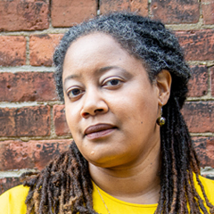Headshot of NK Jemisin: a black woman with dark brown box braids pulled back in a ponytail stands in front of a brick wall wearing a yellow shirt.