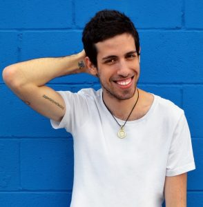 Headshot of Adam, a white man standing in front of a blue wall wearing a white t-shirt and smiles at the camera. His right hand rests on the back of his head.