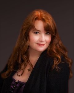 Headshot of Sherrilyn, a white woman with long red hair, wearing a dark blue blouse, sits in front of a black background.