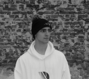 Black and white photo of EDM artist Danilo Kyzyk. He wears a white hoodie and beanie and stands in front of a brick wall looking off into the distance.
