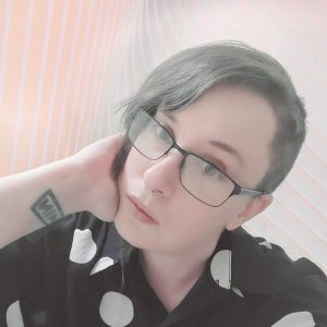Headshot of Kira Leigh: a white transmasc person wearing a black/white shirt and glasses. Their chin rests on their right hand. They are the author of Constellis Voss.