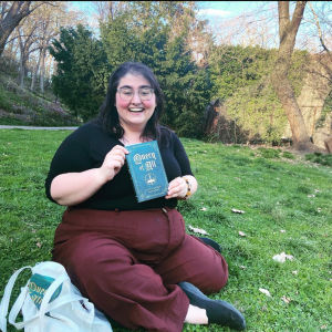 Anya Josephs is a plus-sized white woman wearing a black shirt and maroon pants. She holds her book, Queen Of All, in her hands and smiles at the camera.